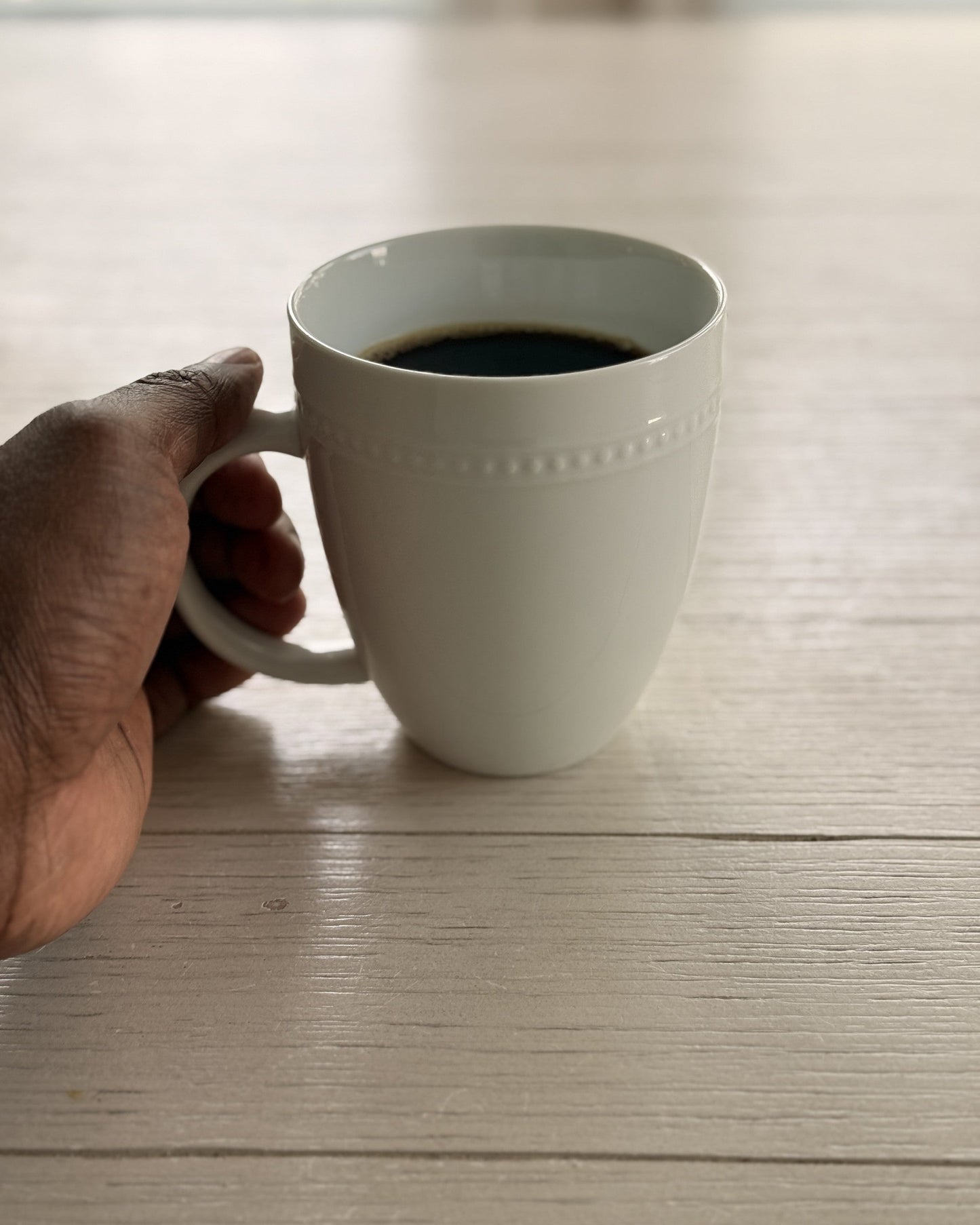a white mug of black coffee held in one hand on a pale wood table