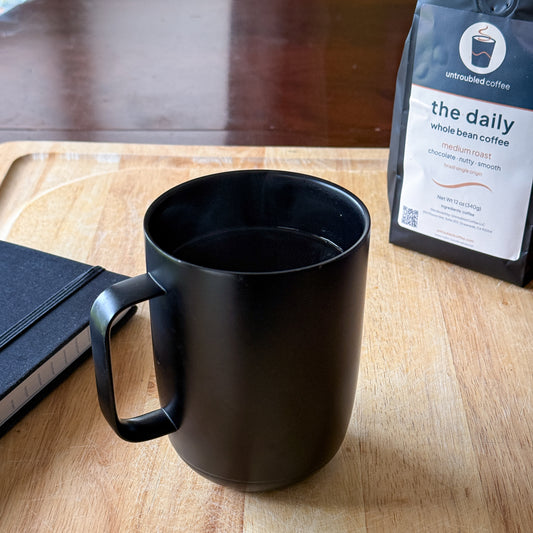 The Daily Brazil single origin medium roast whole bean coffee in a matte black mug on a wood surface, beside a closed journal and bag of coffee.